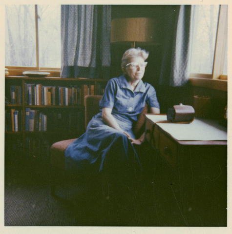 A woman (Lorine Niedecker) sits in a chair looking out the window. In front of her is a desk and behind her is a bookshelf full of books. 