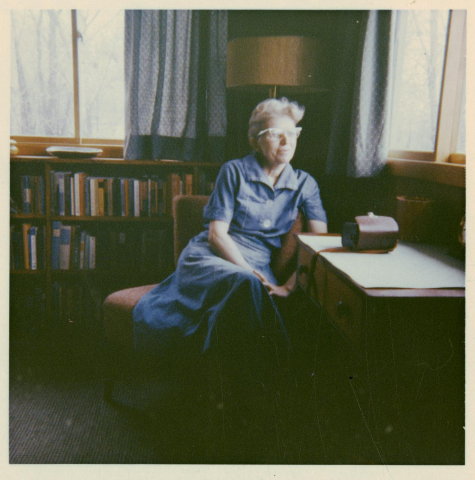 A woman (Lorine Niedecker) sits in a chair looking out the window. In front of her is a desk and behind her is a bookshelf full of books.